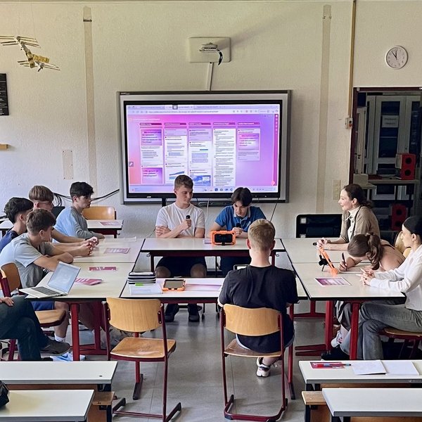 Elf Personen sitzen an zu Kreis zusammengestellten Tischen in einem Klassenraum.