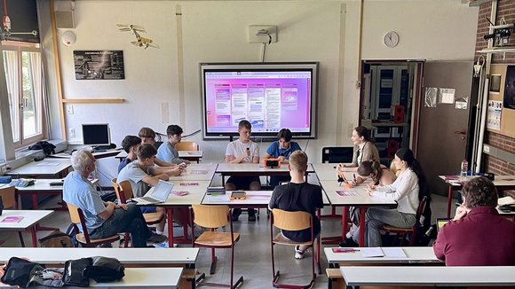 Elf Personen sitzen an zu Kreis zusammengestellten Tischen in einem Klassenraum.