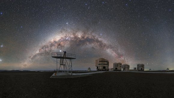 Milchstraße über den vier Teleskopen des Very Large Telescope, davor steht eine Person auf einem Gerüst.