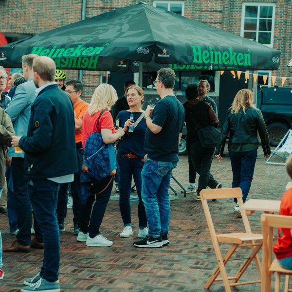 Zahlreiche Menschen stehen auf einem Straßenfest vor Sonnenschirmen und unterhalten sich.