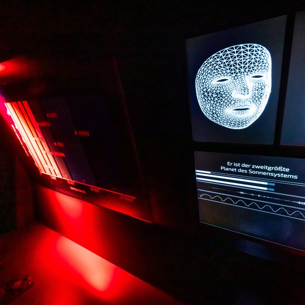 Ein Display zeigt eine weiße Maske, der Raum ist dunkel und nur links rot beleuchtet.