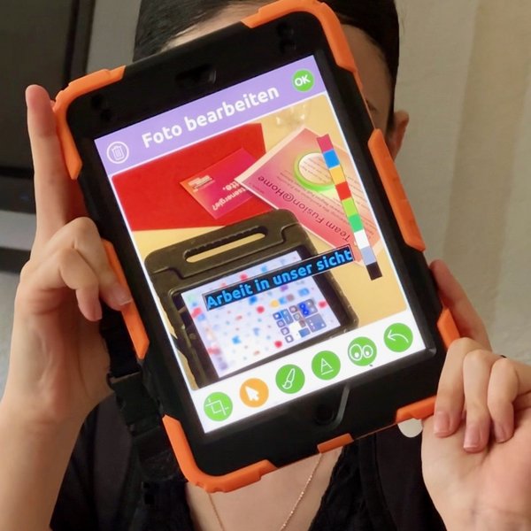 Eine Person hält ein Tablet vor ihr Gesicht, sodass sie nicht zu erkennen ist.