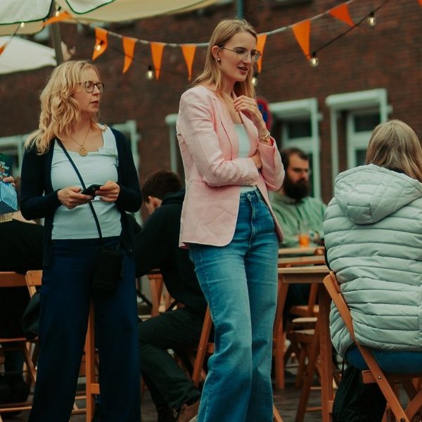 Mehrere Personen sitzen an Tischen unter Sonnenschirmen, drei Personen stehen vor den Tischen.