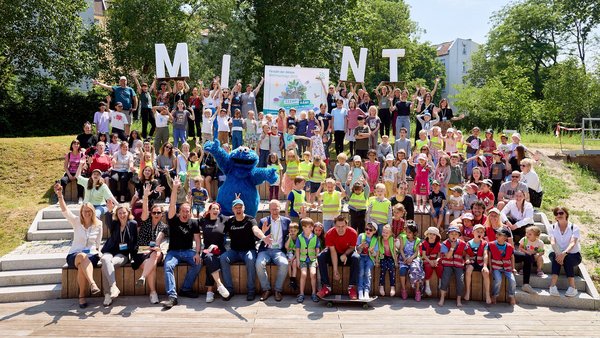 Gruppenbild mit zahlreichen Erwachsenen, Kindern und dem Krümelmonster, die auf auf einer Treppe in einem Park sitzen.