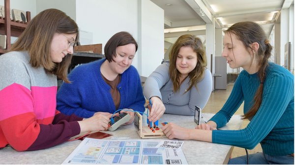 Vier junge Frauen sitzen an einem Tisch in einem hellen Raum und arbeiten gemeinsam an einem Technikprojekt. Sie nutzen ein Messgerät, Kabel und eine kleine Experimentier-Plattform mit Steckmodulen. Auf dem Tisch liegen zudem Informationsblätter mit Diagrammen und QR-Codes.