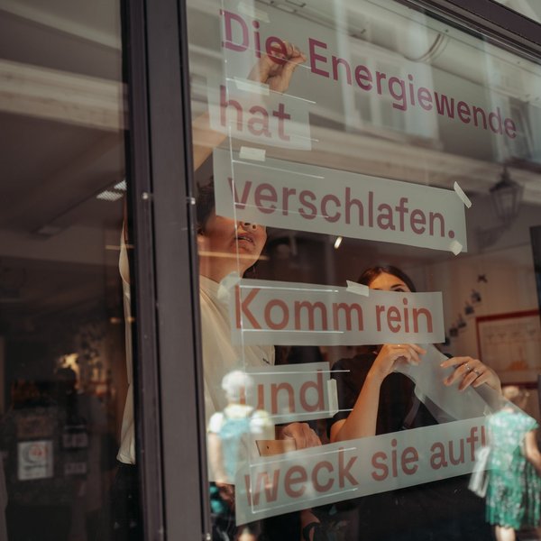 Ein Schaufenster, in welchem von innen zwei Frauen den Text "Die Energiewende hat verschlafen. Komm rein und weck sie auf." aufhängen.