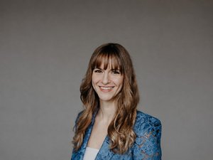 Porträt von Beatrice Aretz: Frau mit langen braunen Haaren und Pony, lächelnd, in blauer Spitzenjacke vor grauem Hintergrund.