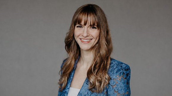 Porträt von Beatrice Aretz: Frau mit langen braunen Haaren und Pony, lächelnd, in blauer Spitzenjacke vor grauem Hintergrund.